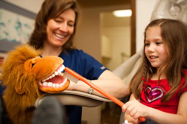 A kid happily playing with a big toothbrush and lion puppet held by Dr. Joy Arend while in a dental chair.